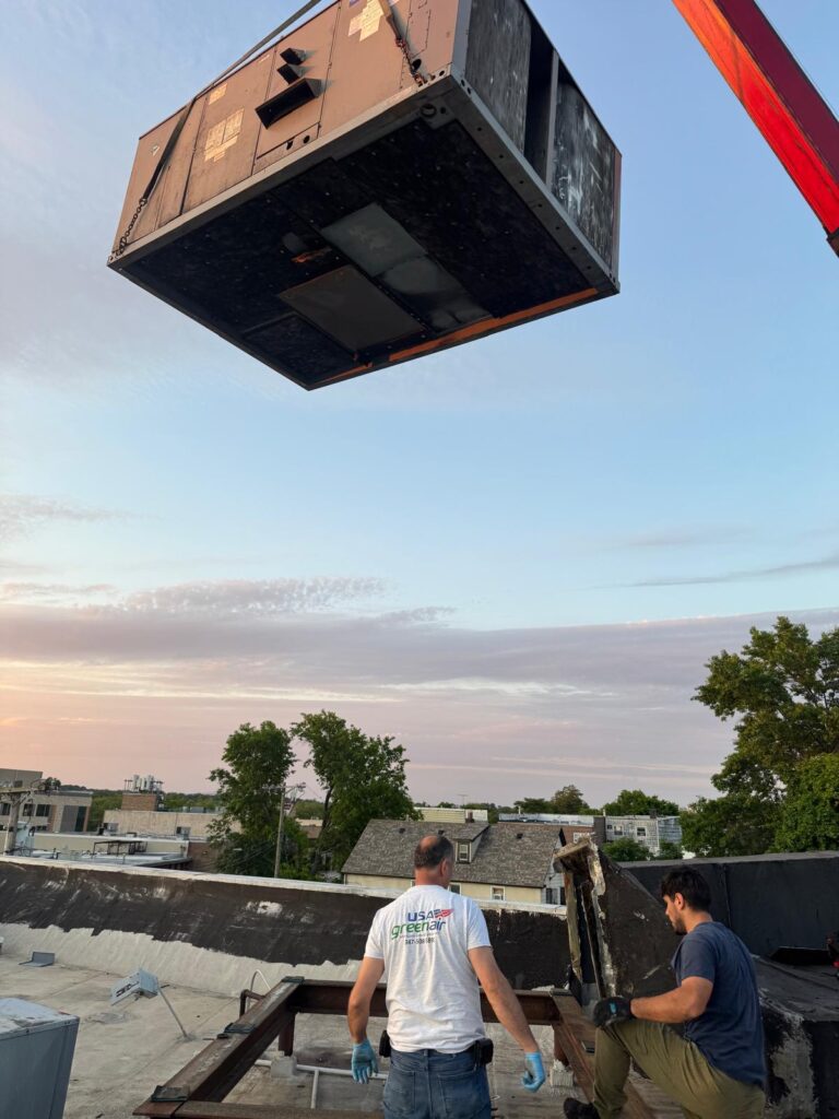USA Green Air technician guiding a rooftop HVAC unit lifted by crane at dusk